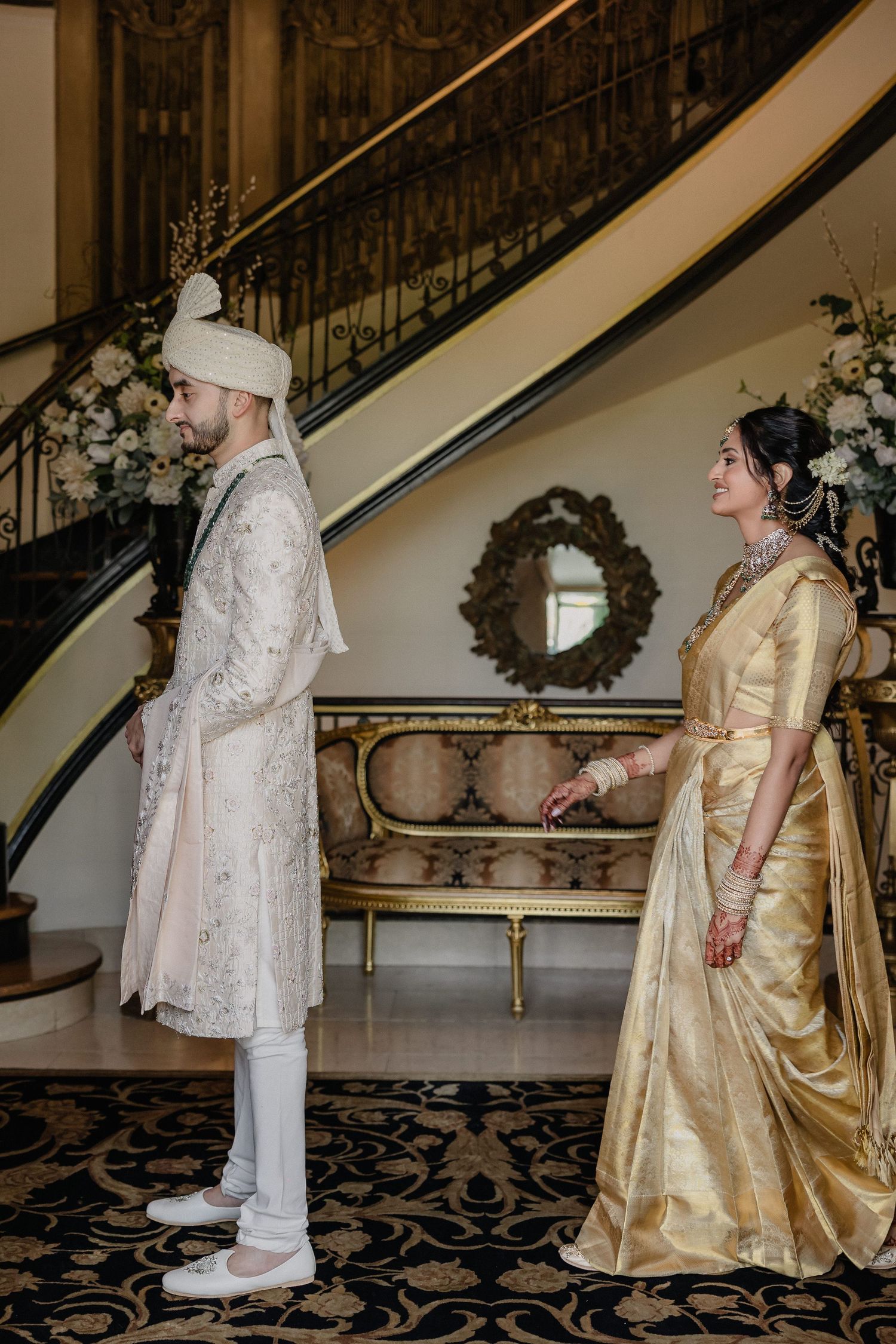 Wedding couple in traditional white and gold attire posing on an elegant staircase with floral decorations.