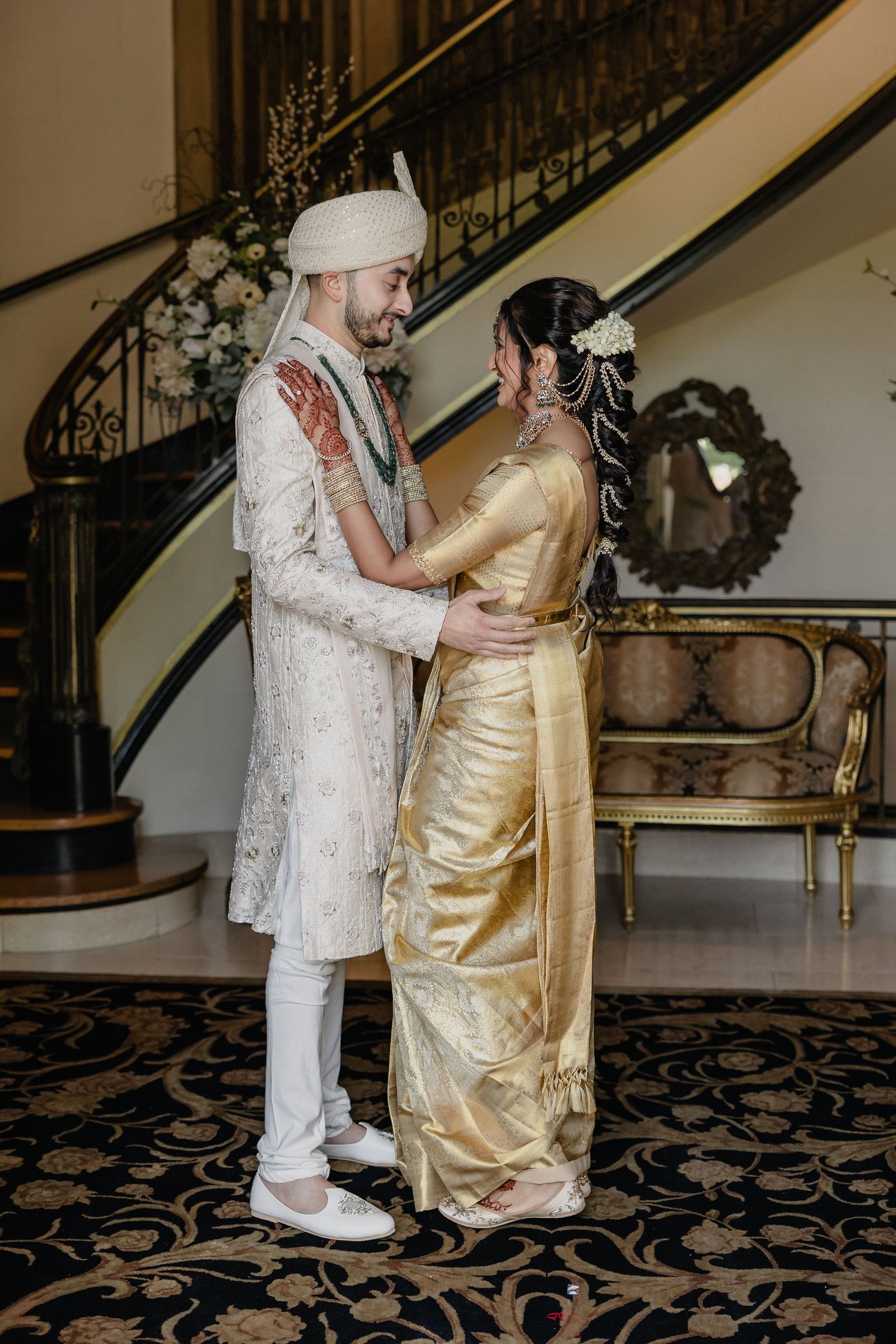 A couple in traditional wedding attire, with the groom in white and bride in gold, share an intimate moment by a staircase.