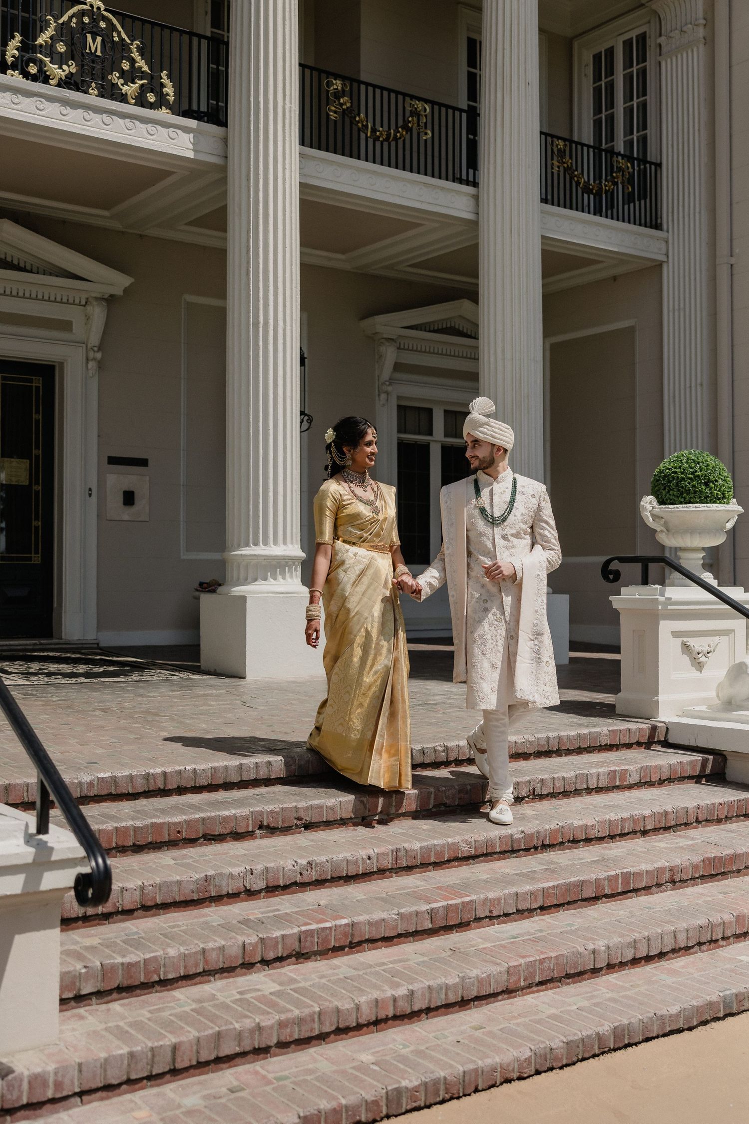 A couple in traditional South Asian wedding attire descend white columned steps of an elegant mansion.