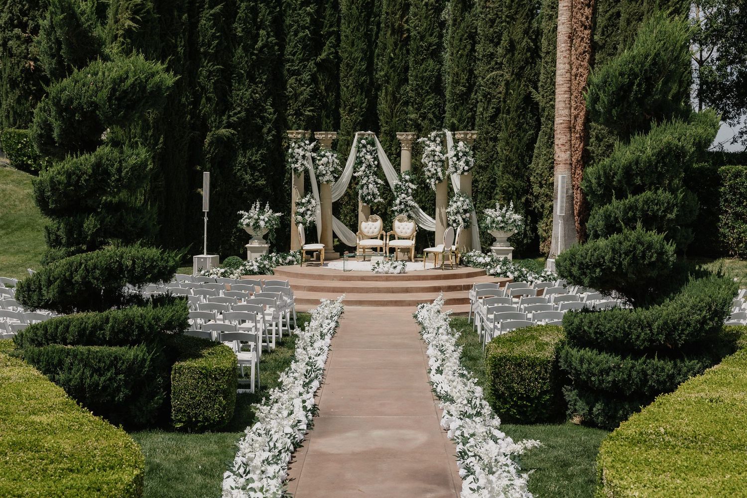 Garden wedding ceremony setup with white floral arch and chairs along a stone pathway.