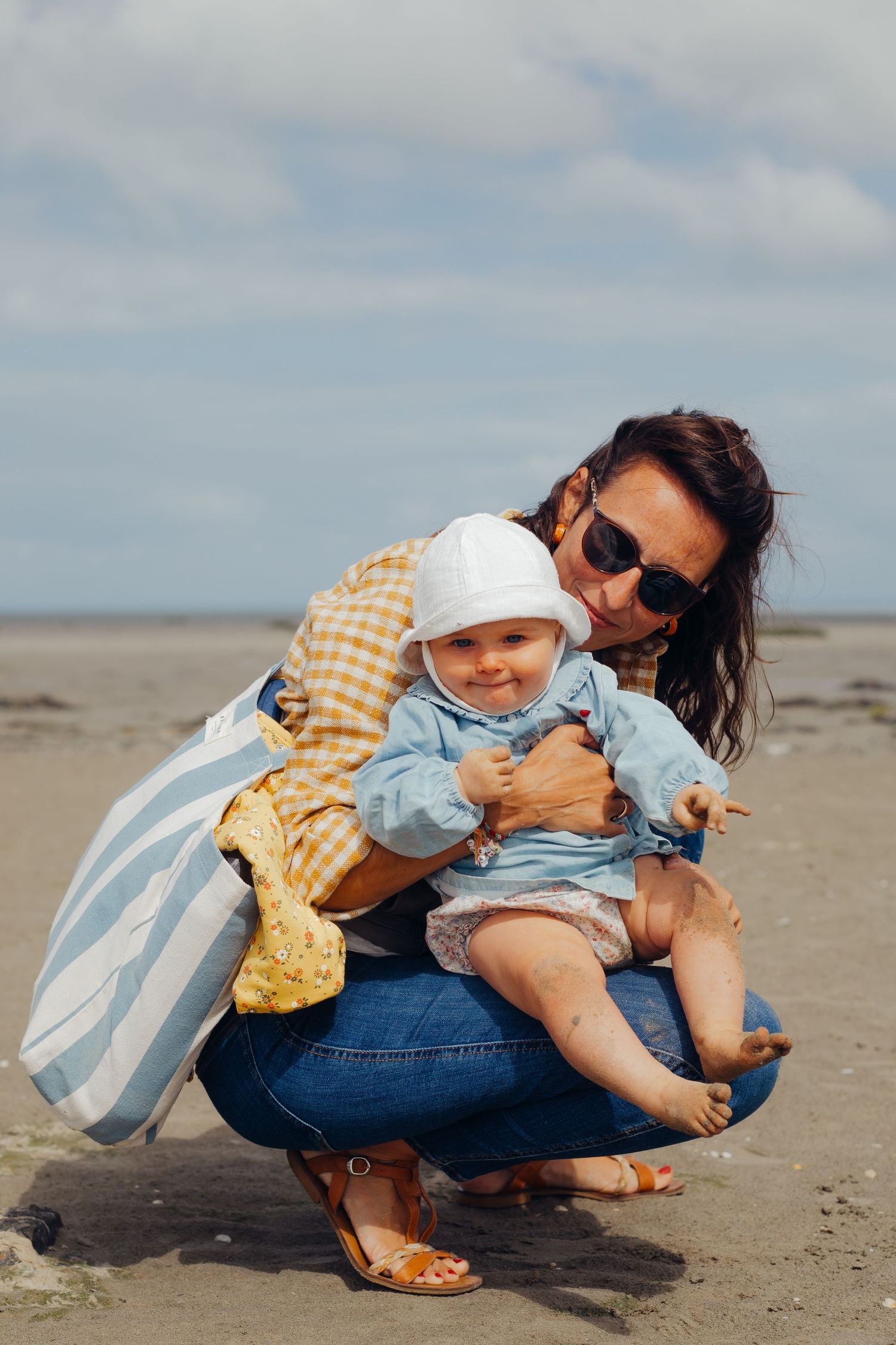 Mother crouching on sandy beach holding smiling baby dressed in blue and white hat.