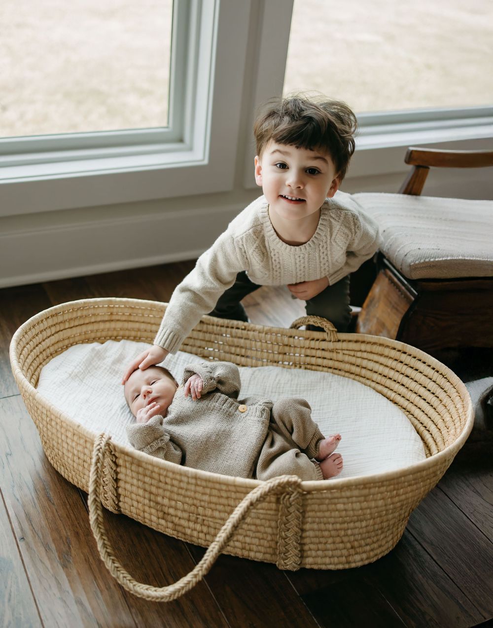 Two children near a window, with a small one resting in a woven Moses basket while another looks on from above.
