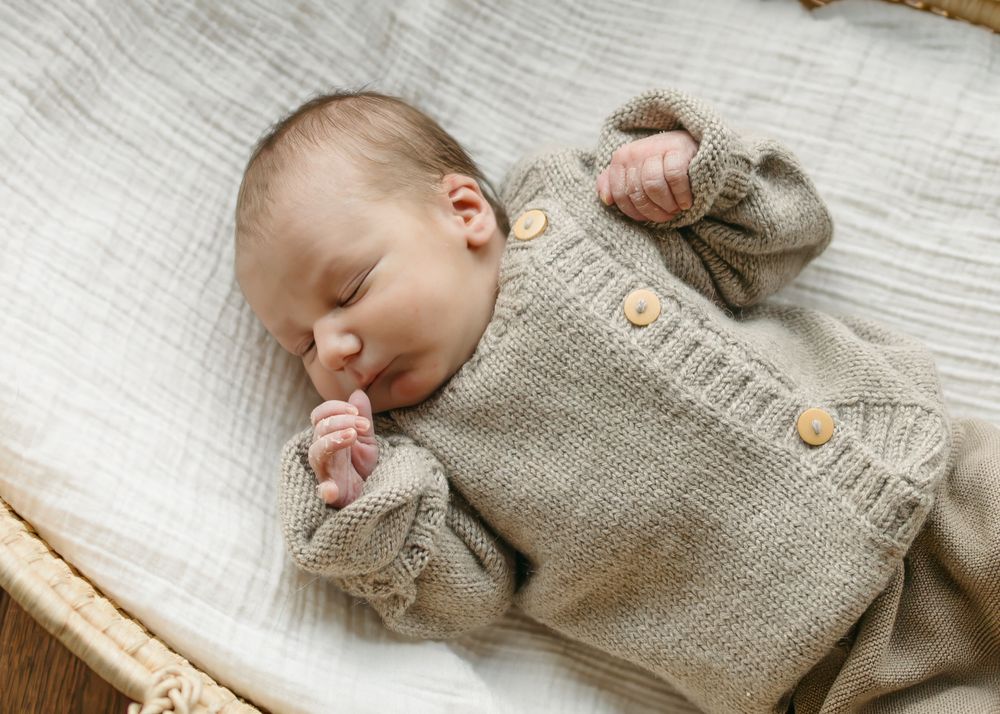 A sleeping baby wearing a cozy beige knitted cardigan sweater with wooden buttons lies peacefully on white bedding.