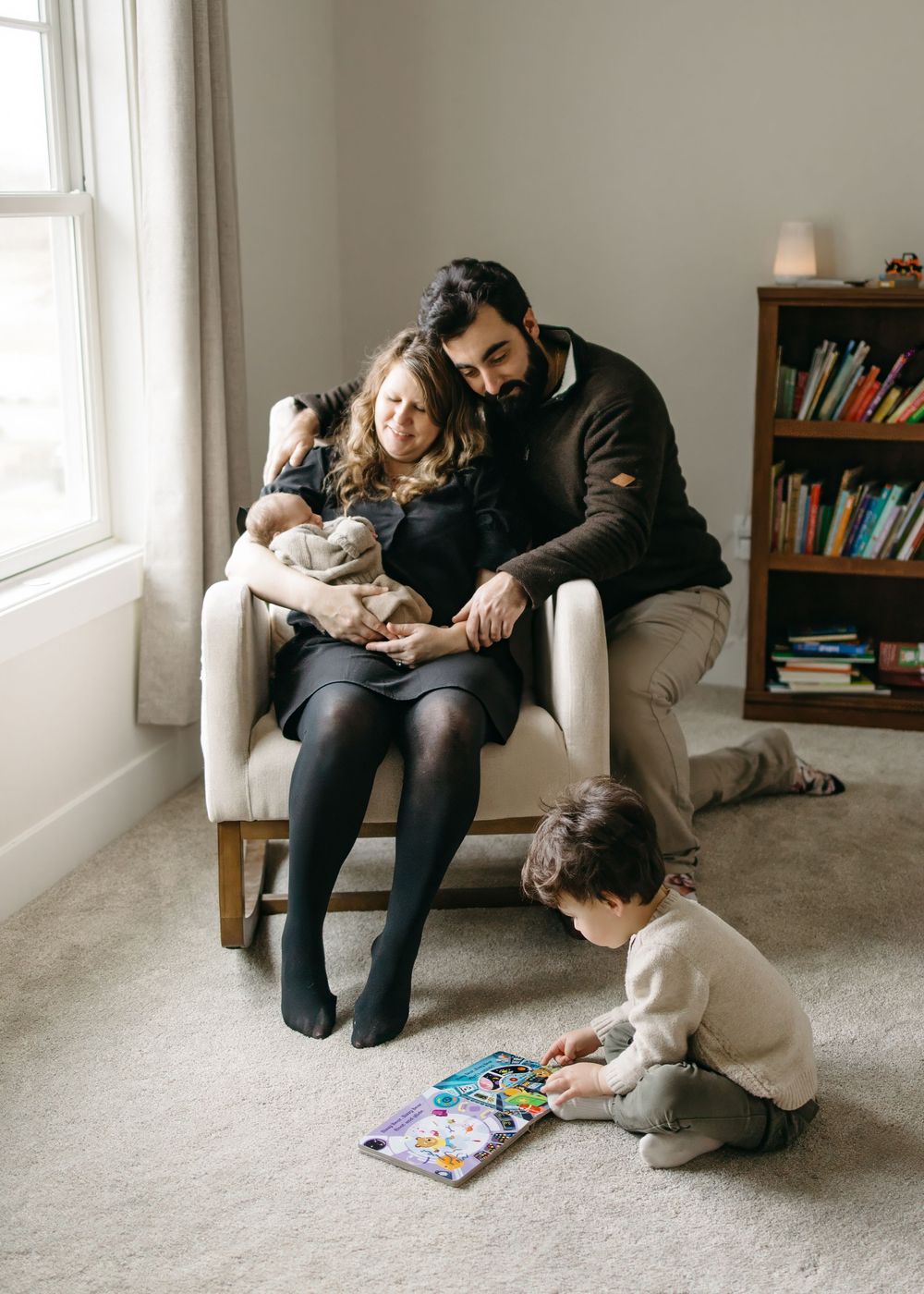 A family sits together on the floor of a cozy room with bookshelves, sharing a quiet moment while reading.