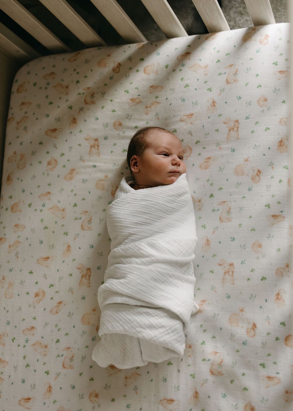 A newborn baby swaddled in white blankets lies on a patterned background showing different stages of swaddling.