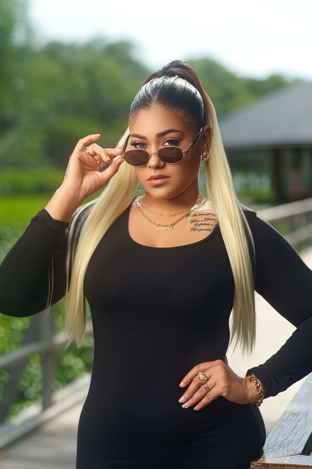 Stylish woman in black outfit and blonde hair poses against scenic outdoor backdrop with sunglasses.