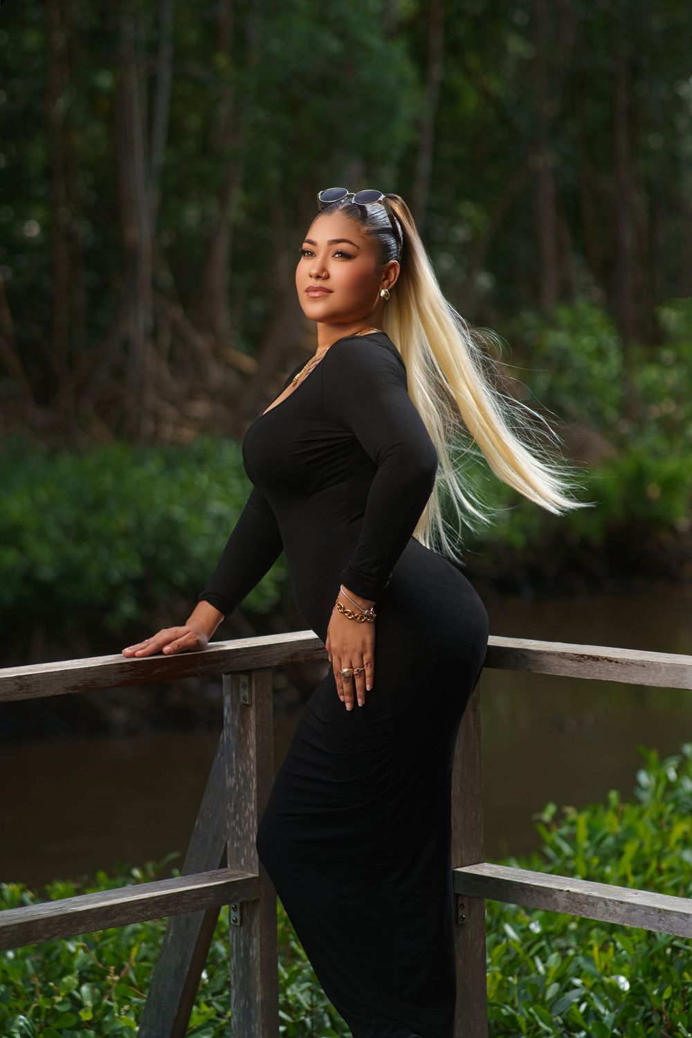 A figure in a form-fitting black dress poses on a wooden deck overlooking a serene waterway.