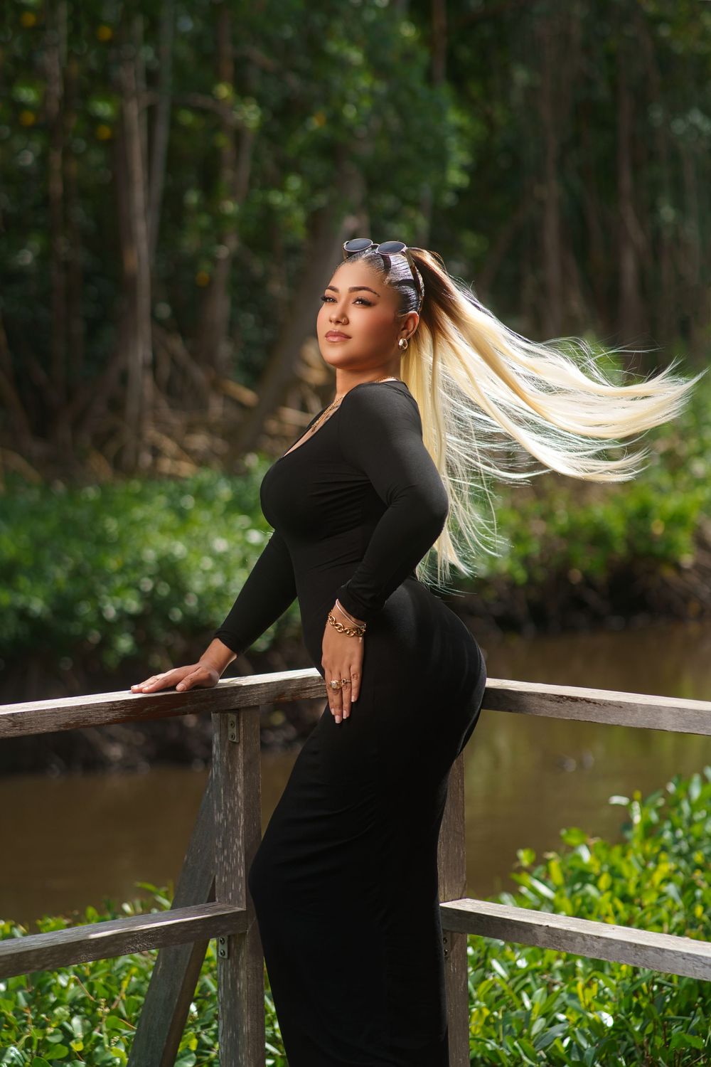 Woman in black dress poses gracefully on wooden deck railing by river in natural outdoor setting.