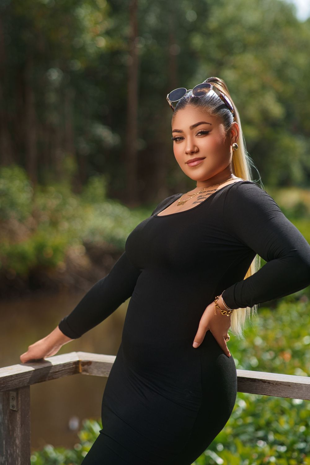 Woman in black long-sleeve dress poses elegantly against wooden railing in outdoor nature setting.