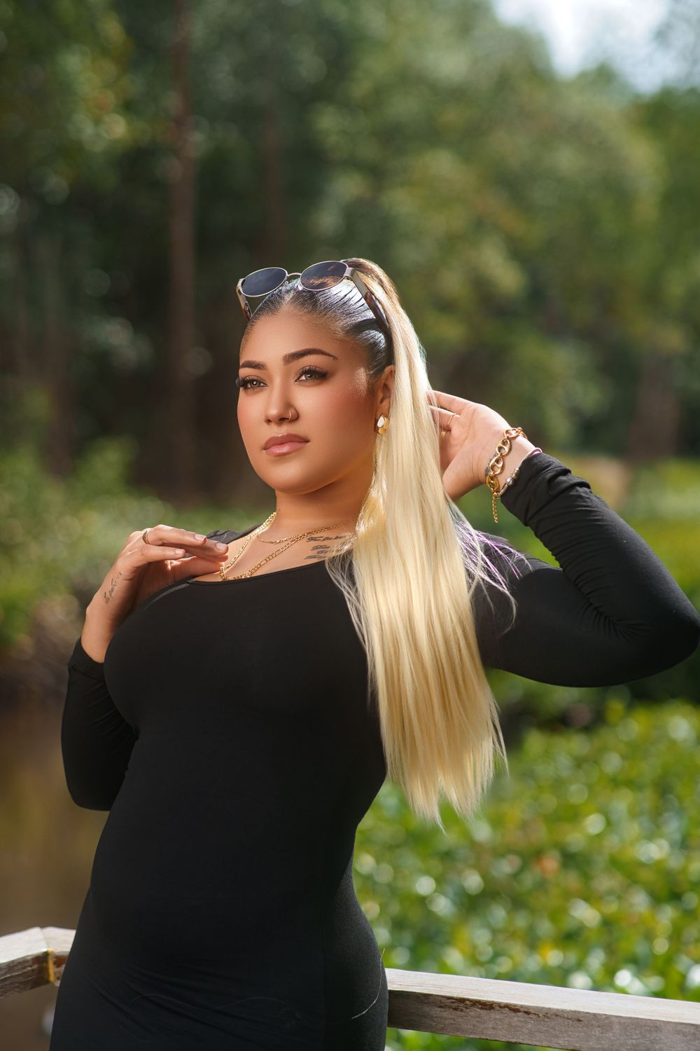 Model in black long sleeve top poses outdoors against green foliage background in fashion photoshoot series.