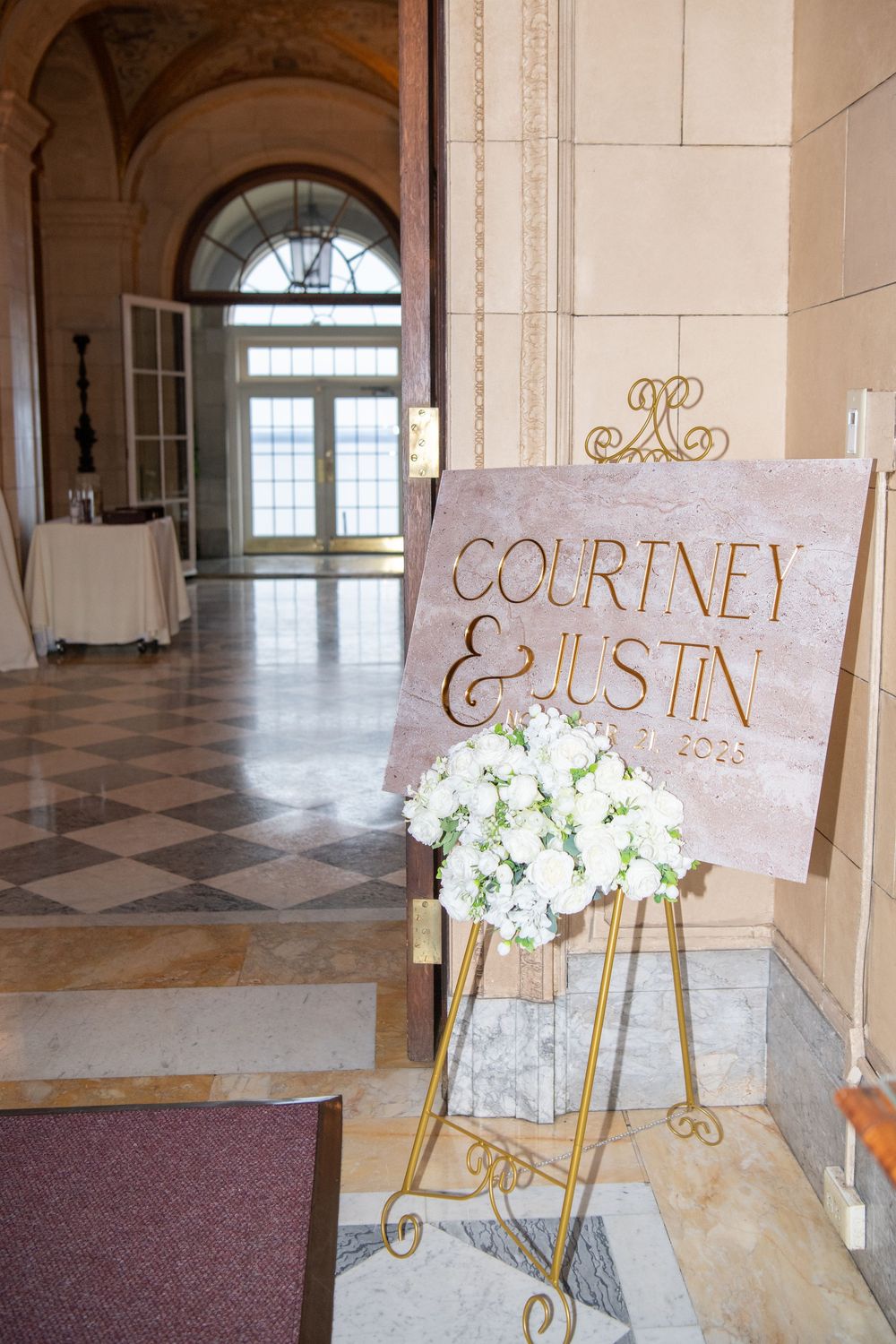 Wedding welcome sign with white floral arrangement on gold easel in elegant hallway.
