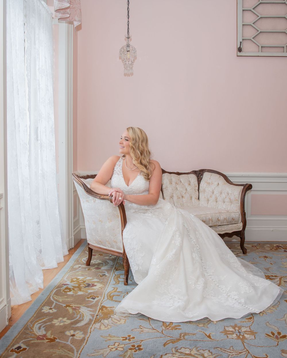 Bride seated on vintage settee by window in pink room with ornate carpet.