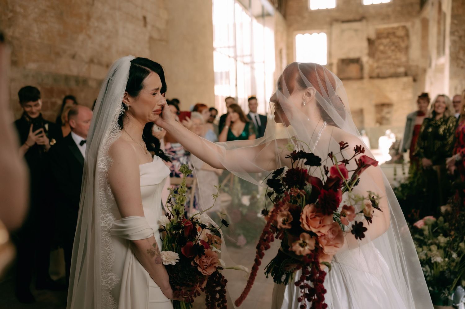 Wedding party members stand in a rustic stone venue with colorful floral bouquets during a ceremony.
