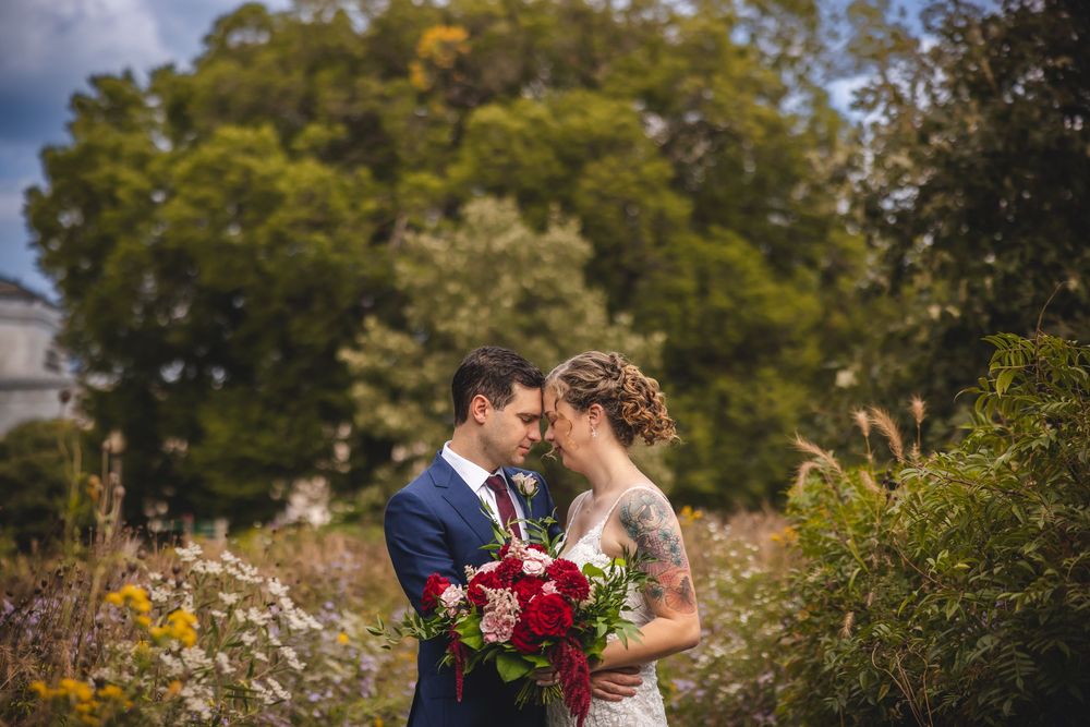 A couple embraces in a romantic garden setting surrounded by lush autumn foliage and blooming flowers.