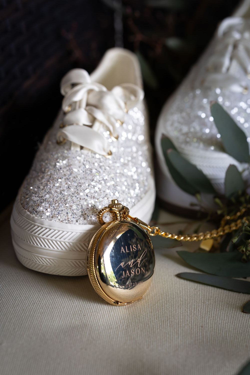 Sparkly white wedding sneaker with gold locket and eucalyptus leaves on light fabric.