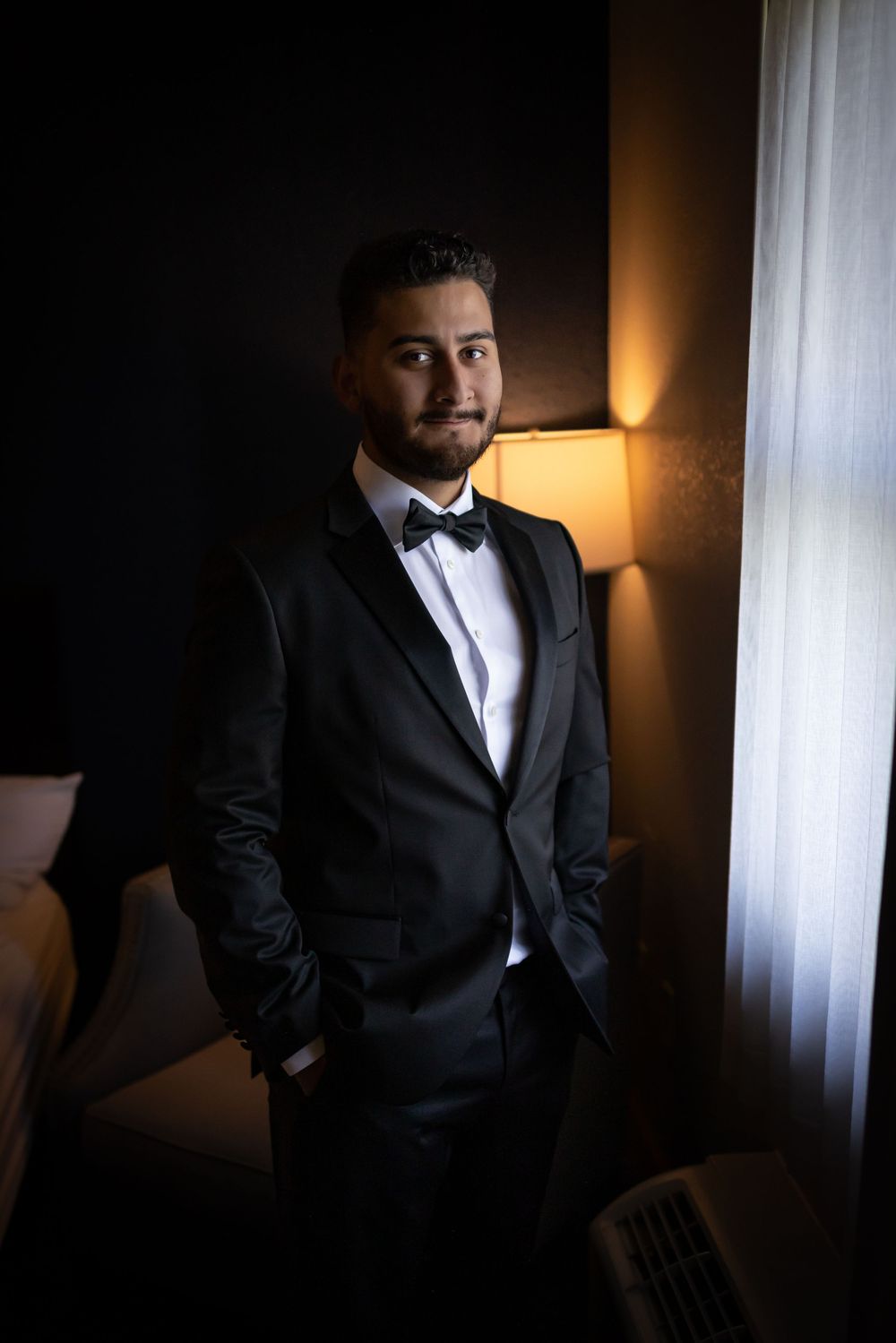 A groom in a black tuxedo poses near a window with dramatic lighting and a warm lamp glow in the background.