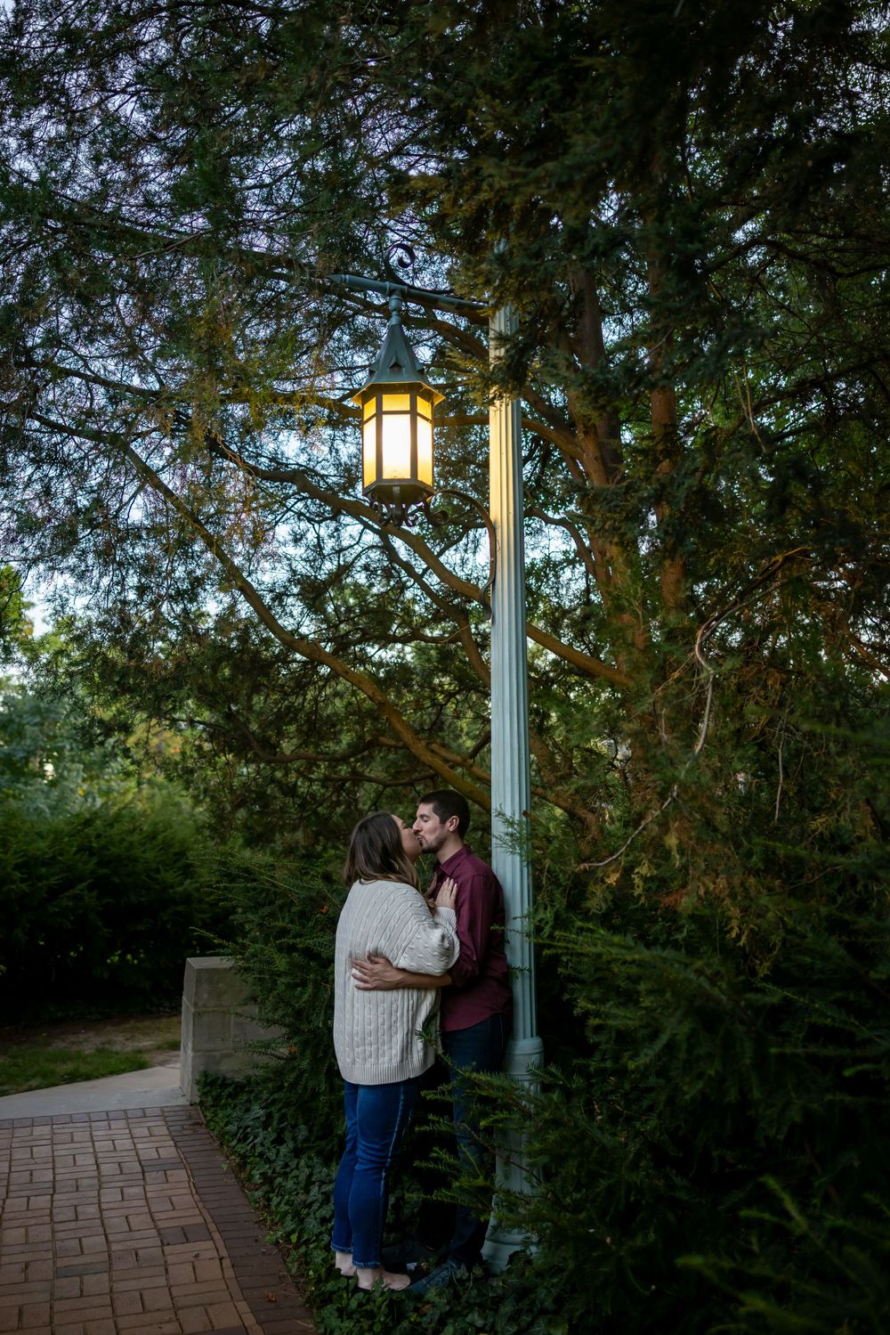 A romantic evening moment under a glowing lamppost on a brick path surrounded by lush greenery.