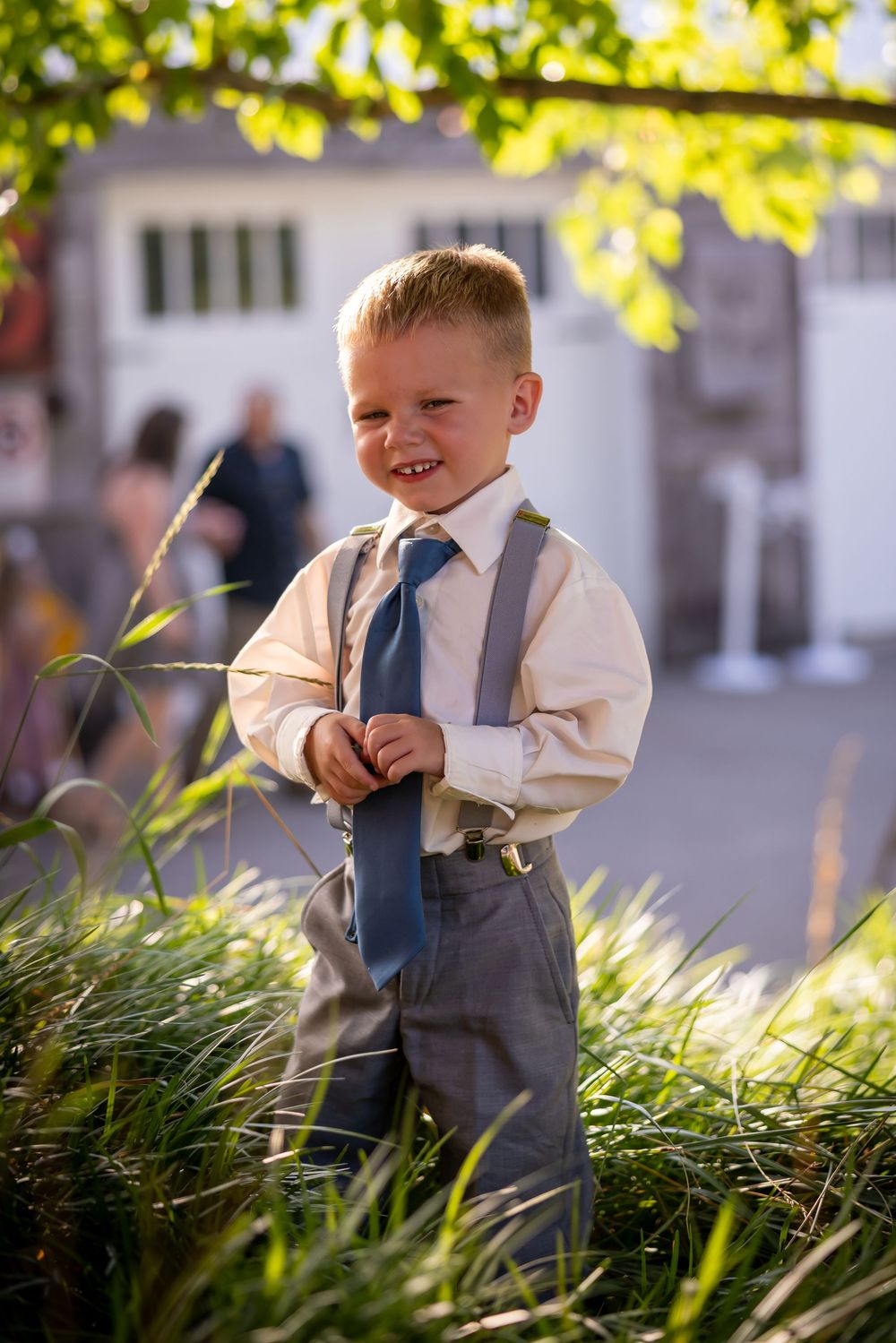 A young ring bearer in dress clothes smiles while standing in garden foliage.