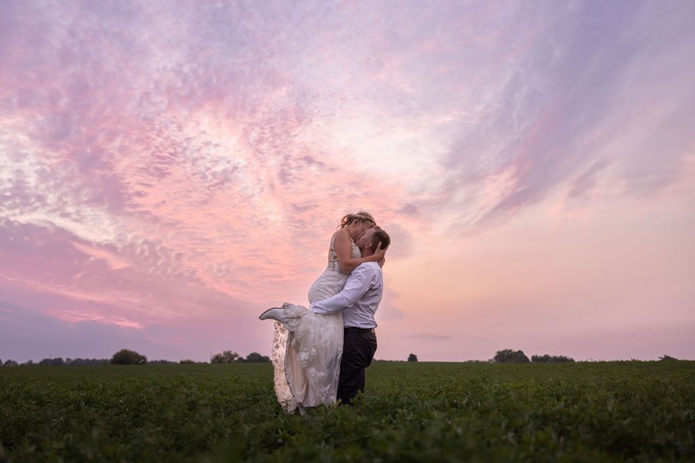 A couple embraces in a grassy field against a dramatic pink and purple sunset sky.