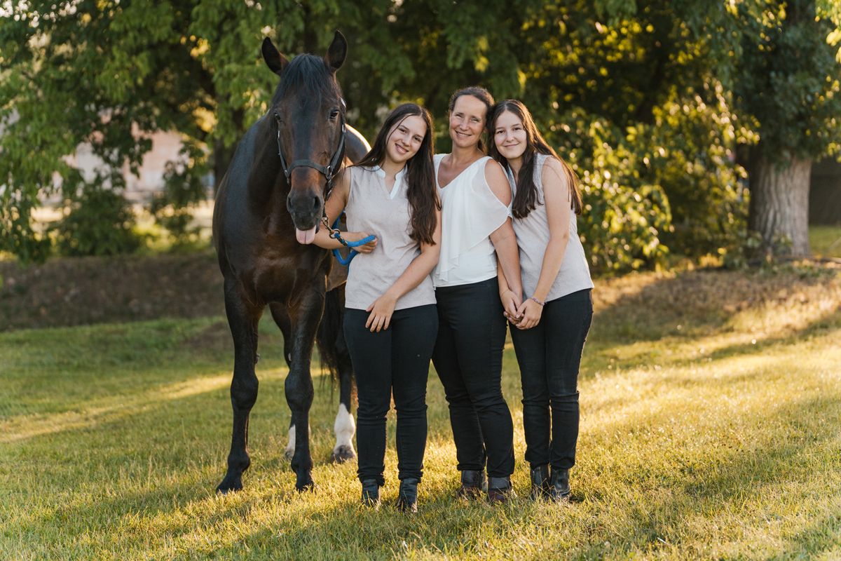 A mom and her daughters in matching black and white outfits poses together on a grassy hill during golden hour with their horse photoshoot