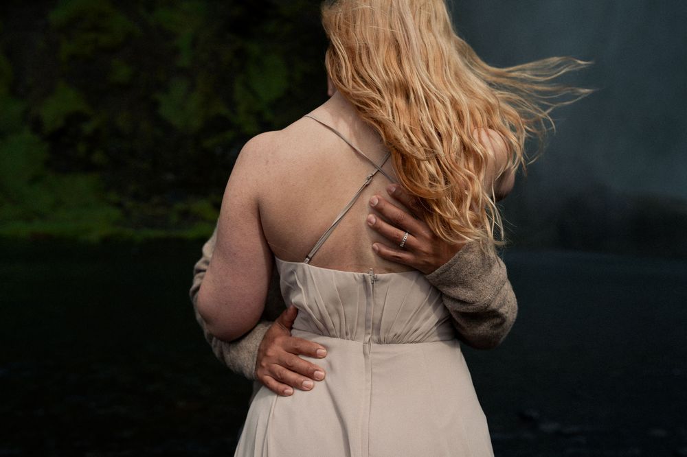 Woman in elegant white dress embracing herself outdoors near dark water.