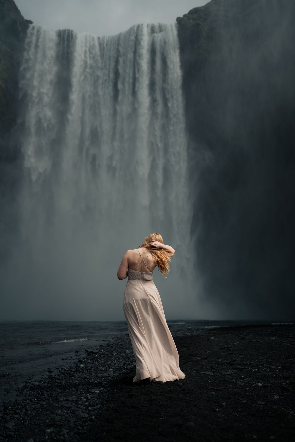 Woman in flowing white gown standing before majestic Skogafoss waterfall in Iceland.