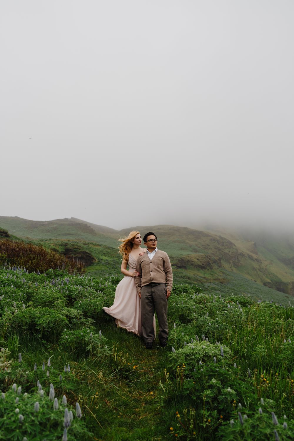 Couple embracing on misty green hillside in Iceland during foggy day.