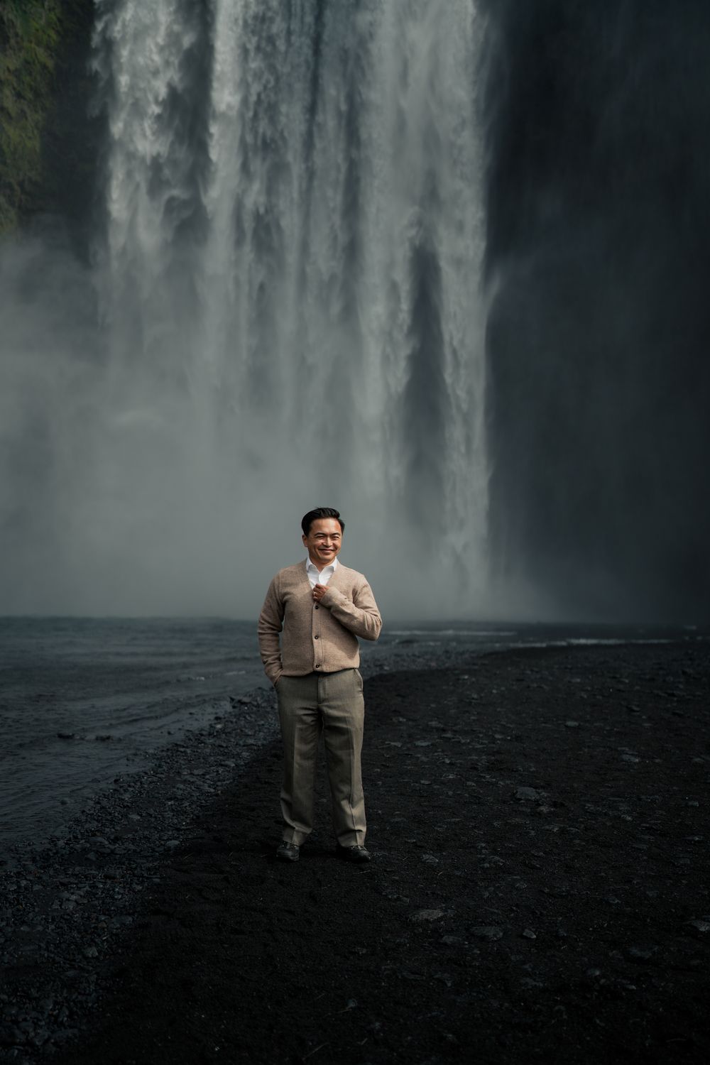 Smiling man in beige suit standing before dramatic Skogafoss waterfall Iceland.