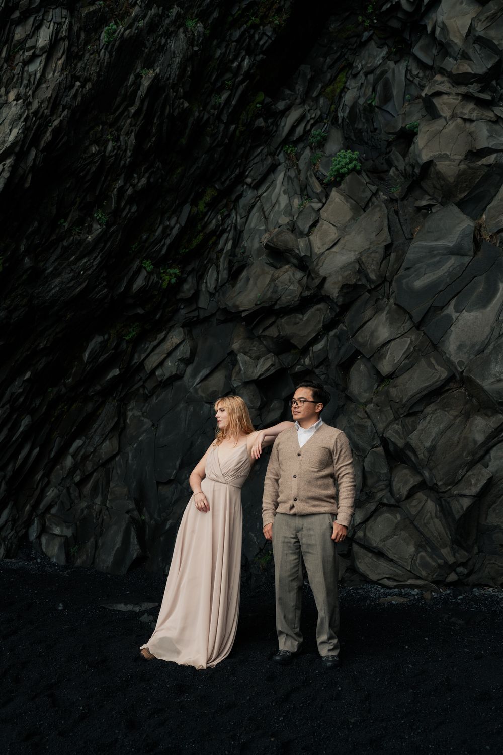 Couple posing against dramatic dark volcanic rock wall on Iceland black beach.