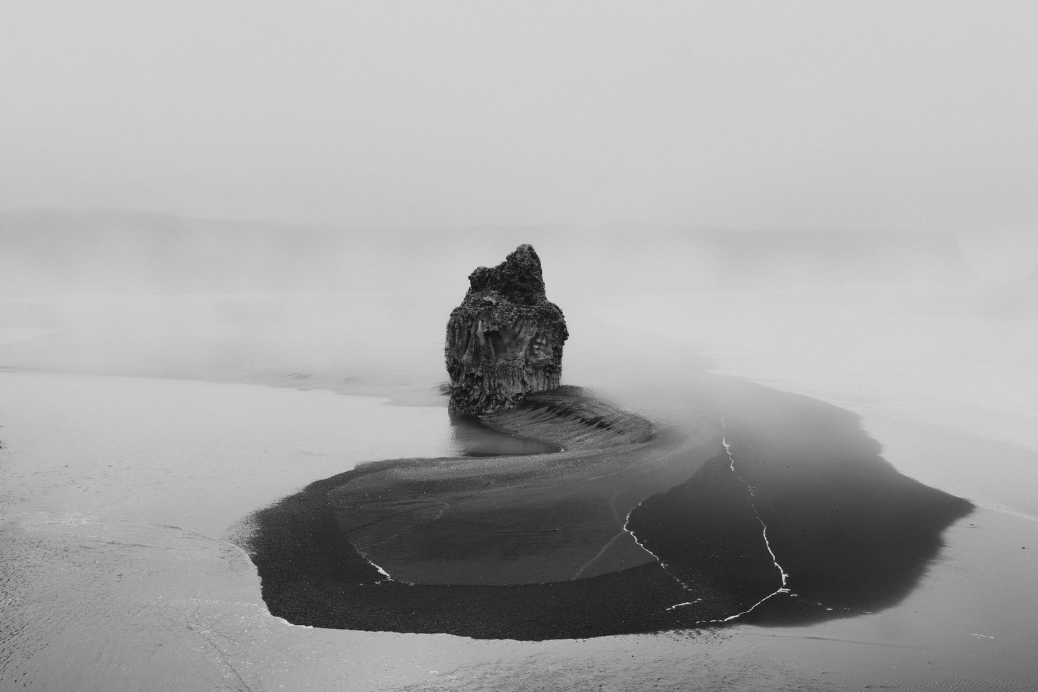 A dramatic black and white photo of a lone rock formation on a misty black sand beach.
