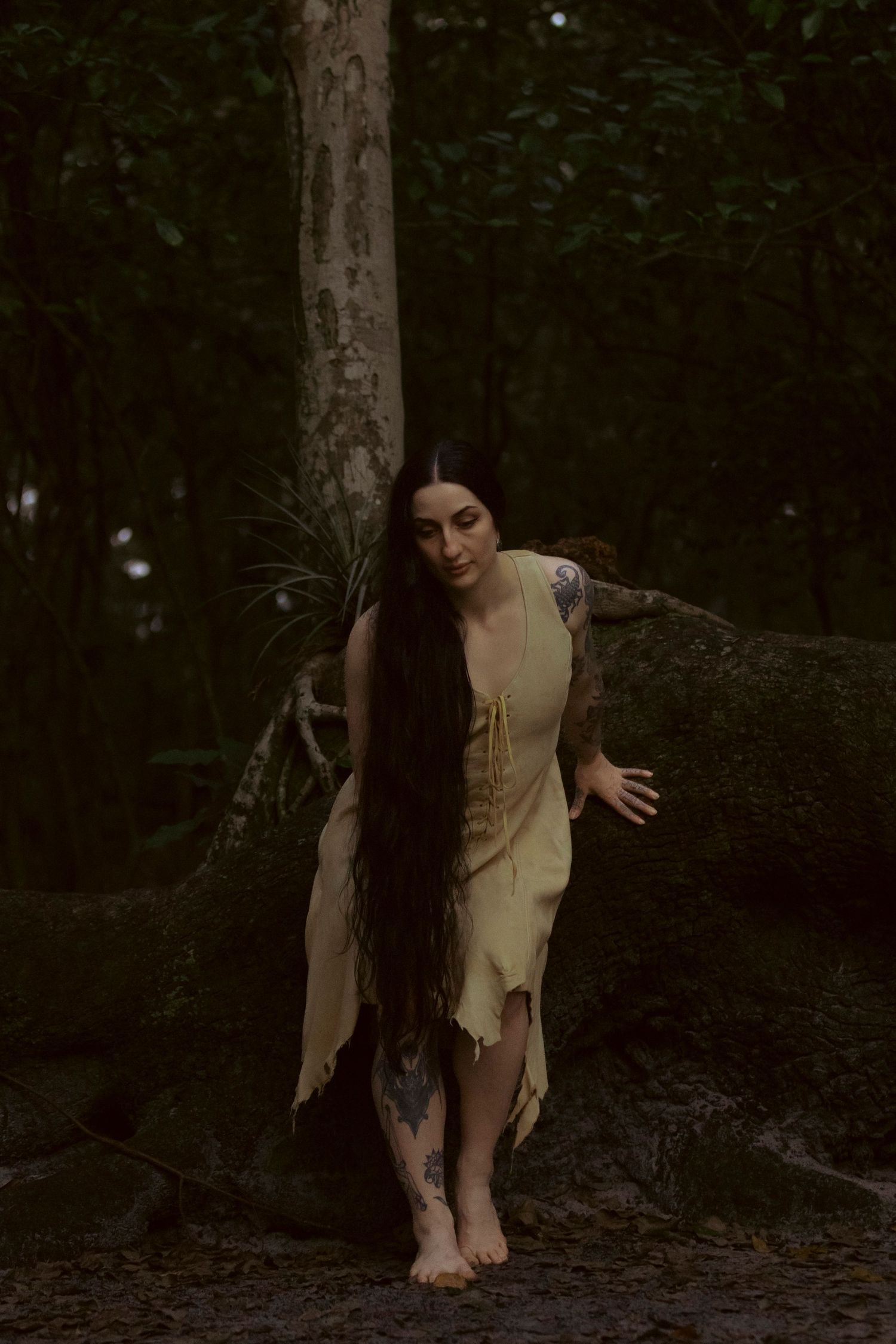 A figure in a tattered beige dress with extremely long dark hair stands barefoot against a tree in a dark forest setting.