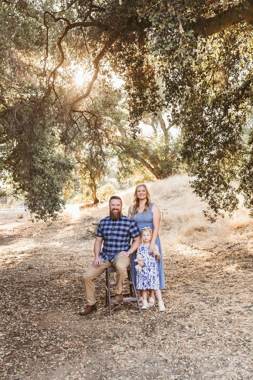 A family poses together on a sunlit dirt path under oak trees during golden hour.