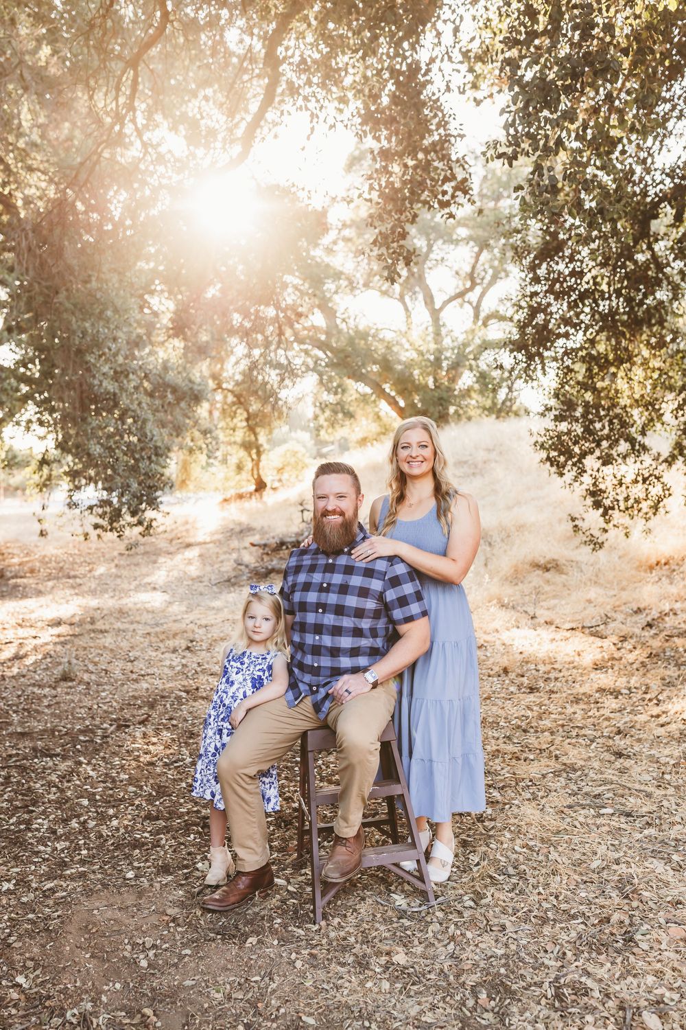 A family sits together on a wooden bench in a sun-dappled outdoor setting during golden hour.