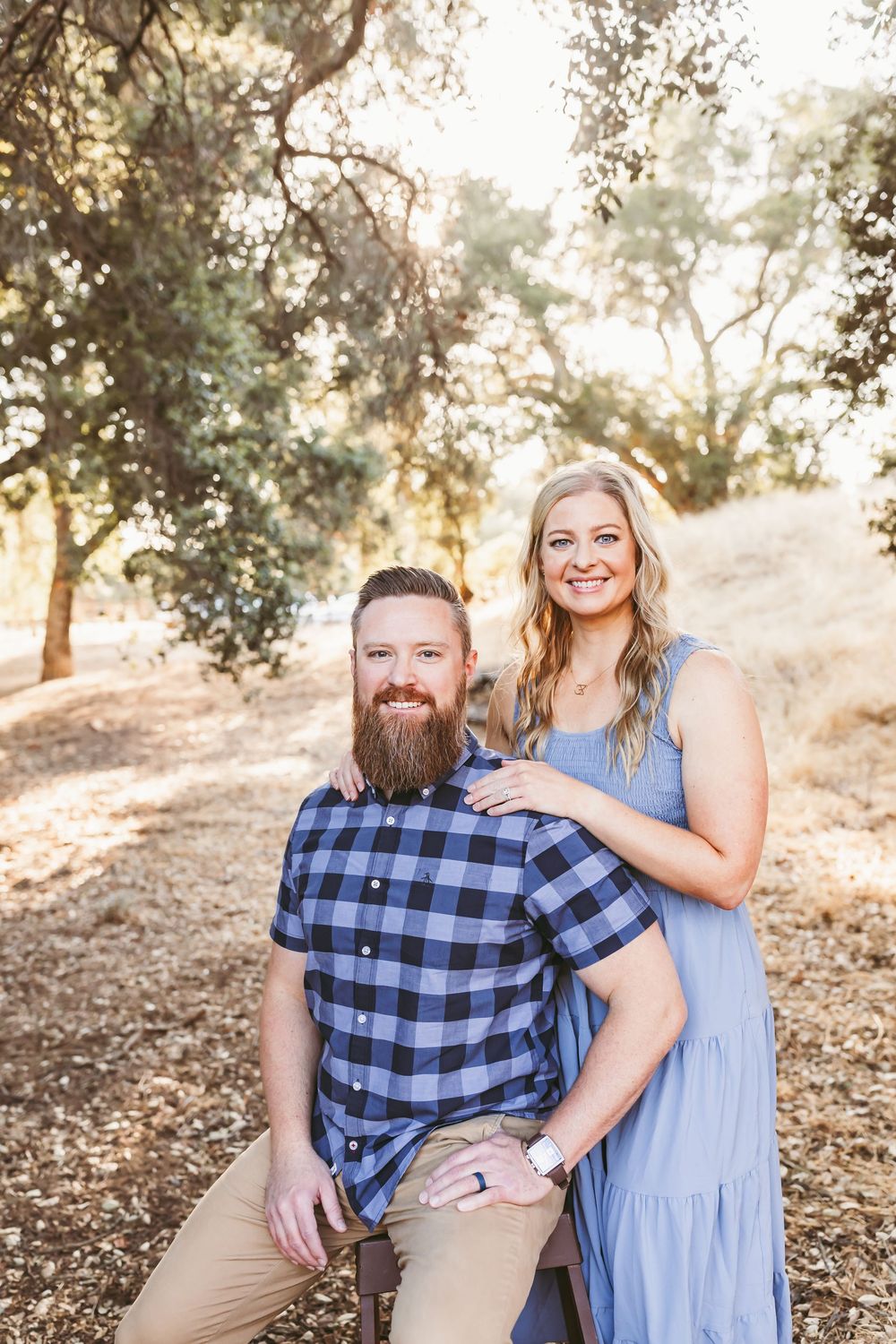 A couple in coordinated blue outfits poses together in a sun-dappled wooded setting during a photo session.