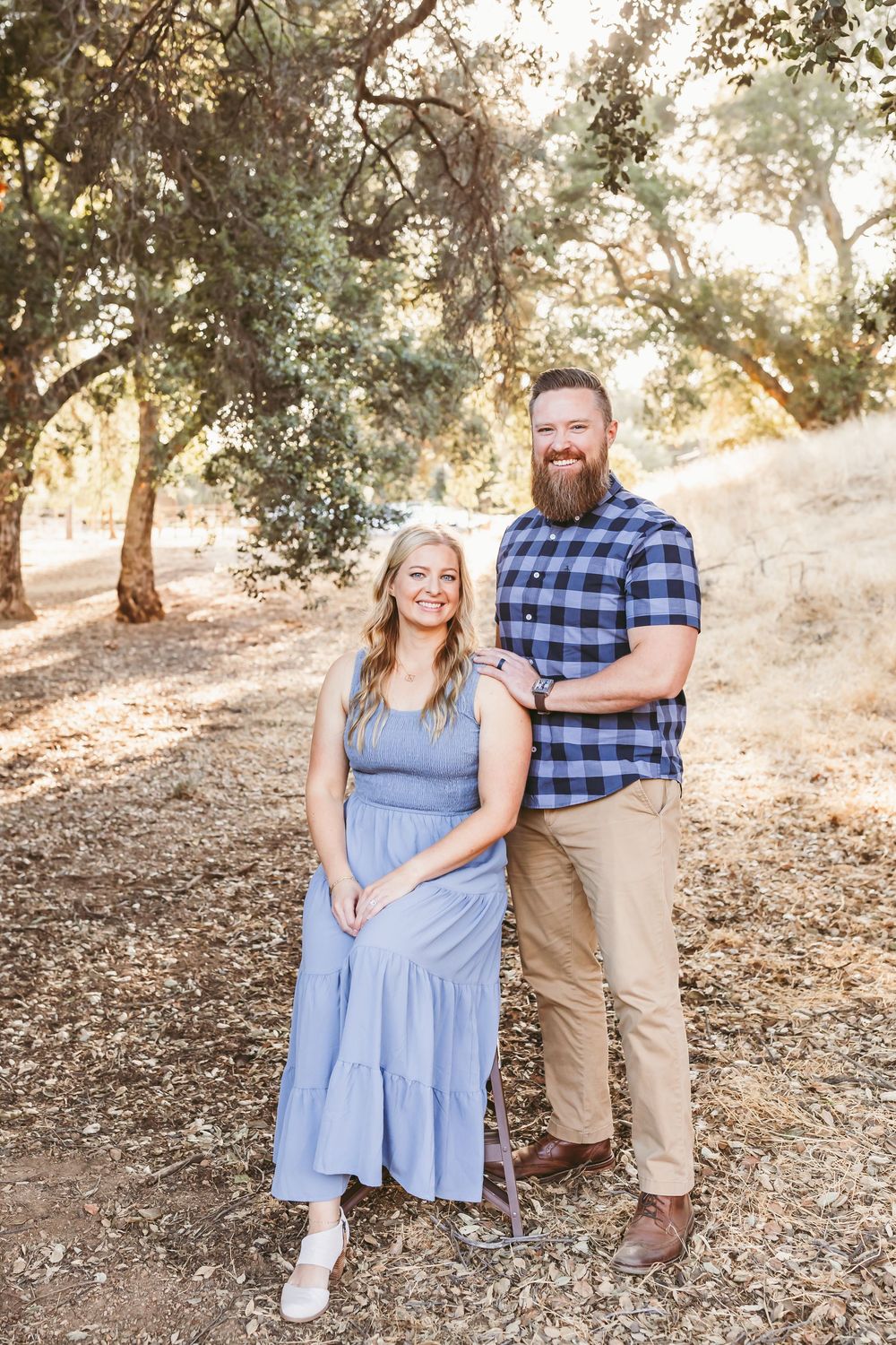 A couple poses together outdoors on a sunny hillside with trees in the background during a casual photo session.