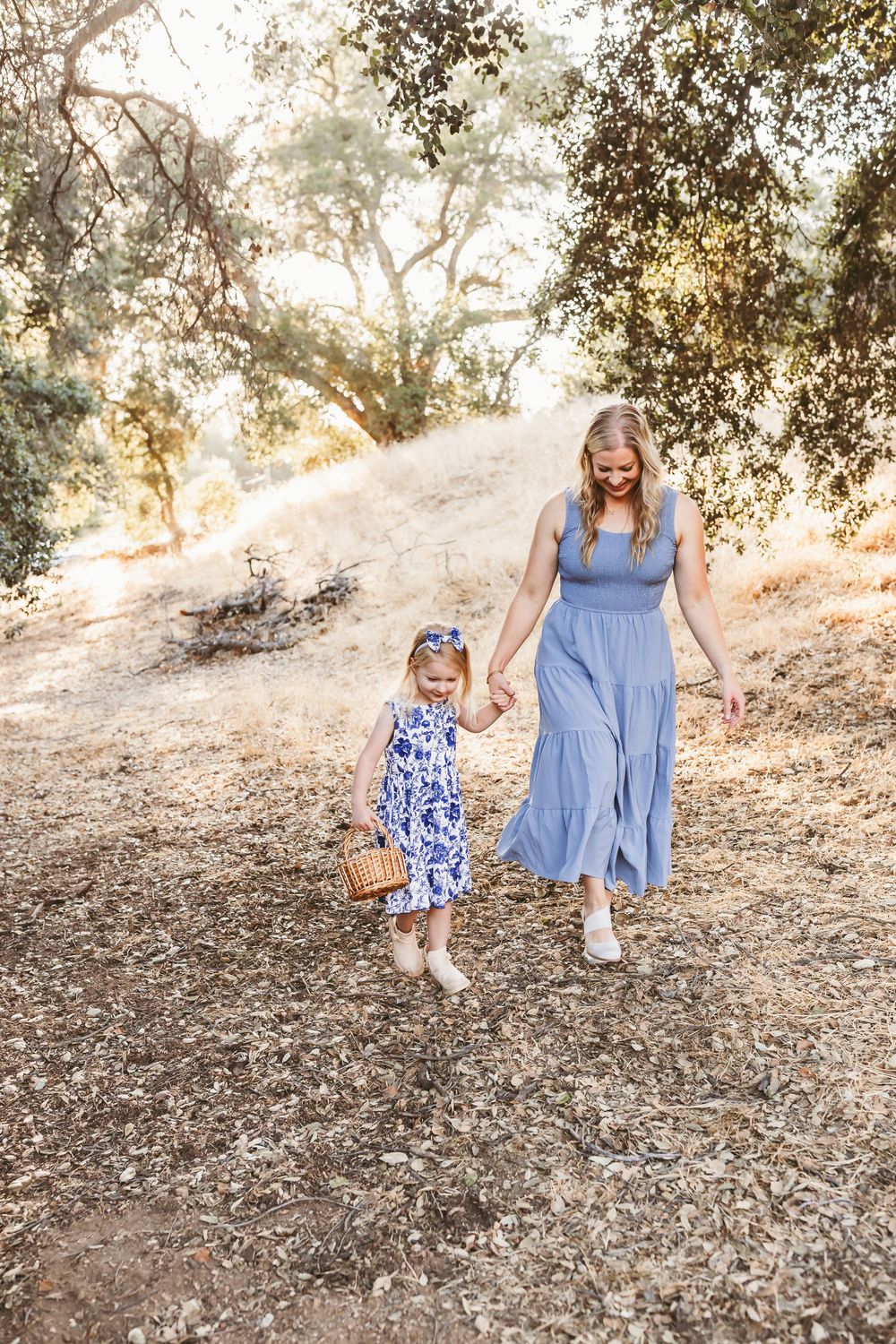 Mother and daughter in blue dresses walk together on a sunlit hillside path during golden hour.