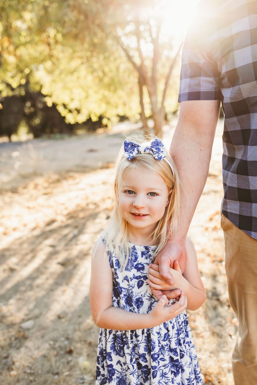 A child in a blue and white floral dress smiles while holding hands outdoors in golden sunlight.