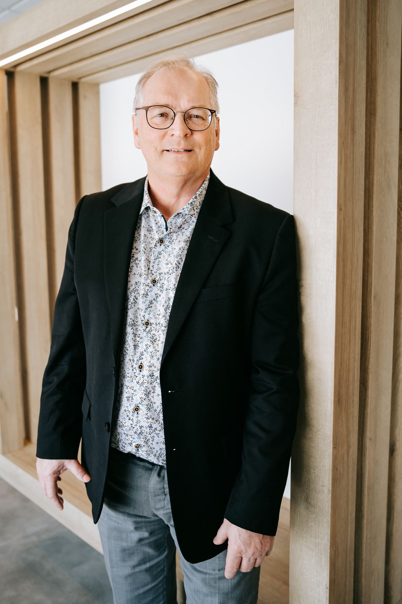 Professional portrait of a person wearing a black blazer and patterned shirt against a neutral hallway background.