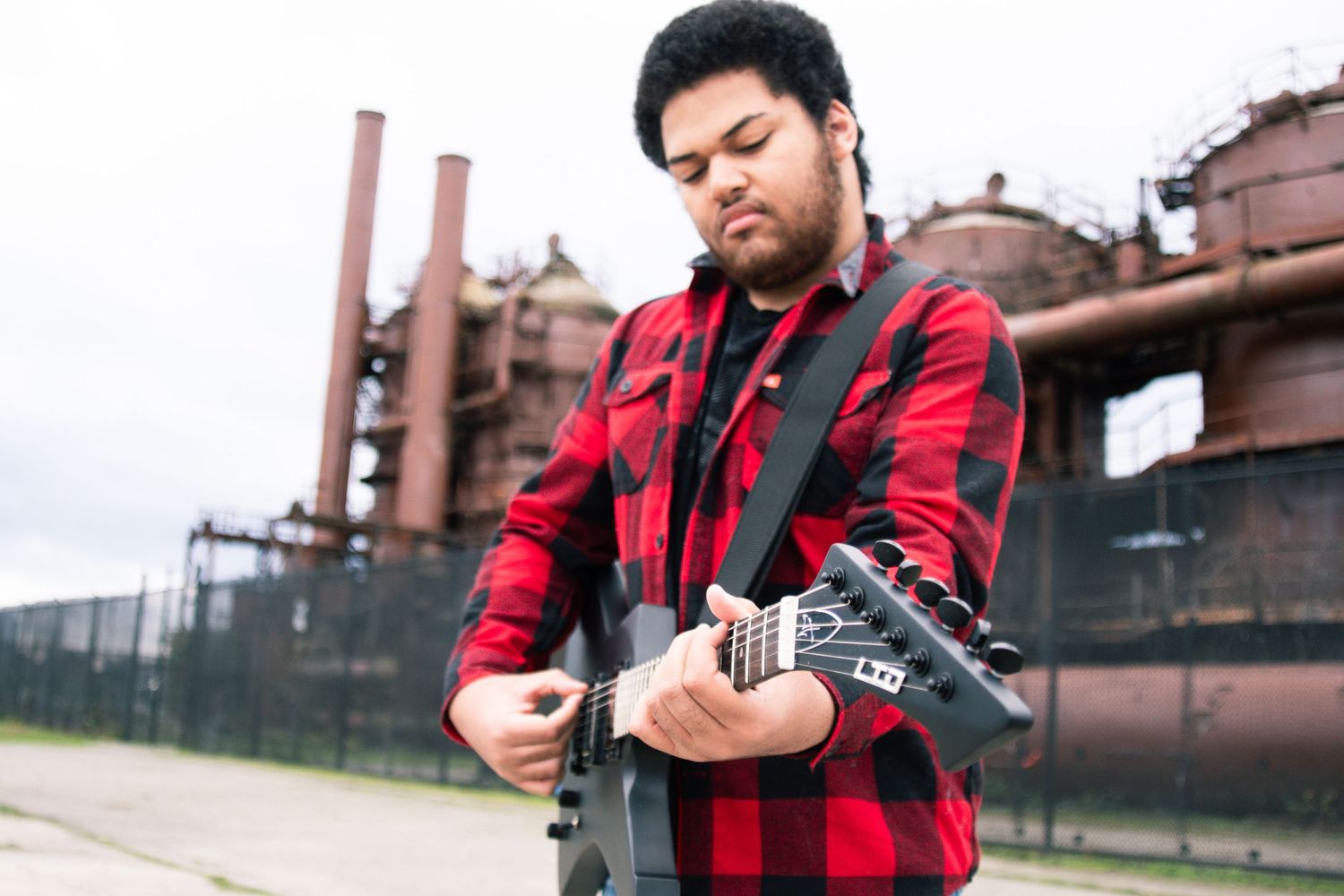 Person in red and black plaid jacket poses in front of industrial buildings in multiple artistic shots.