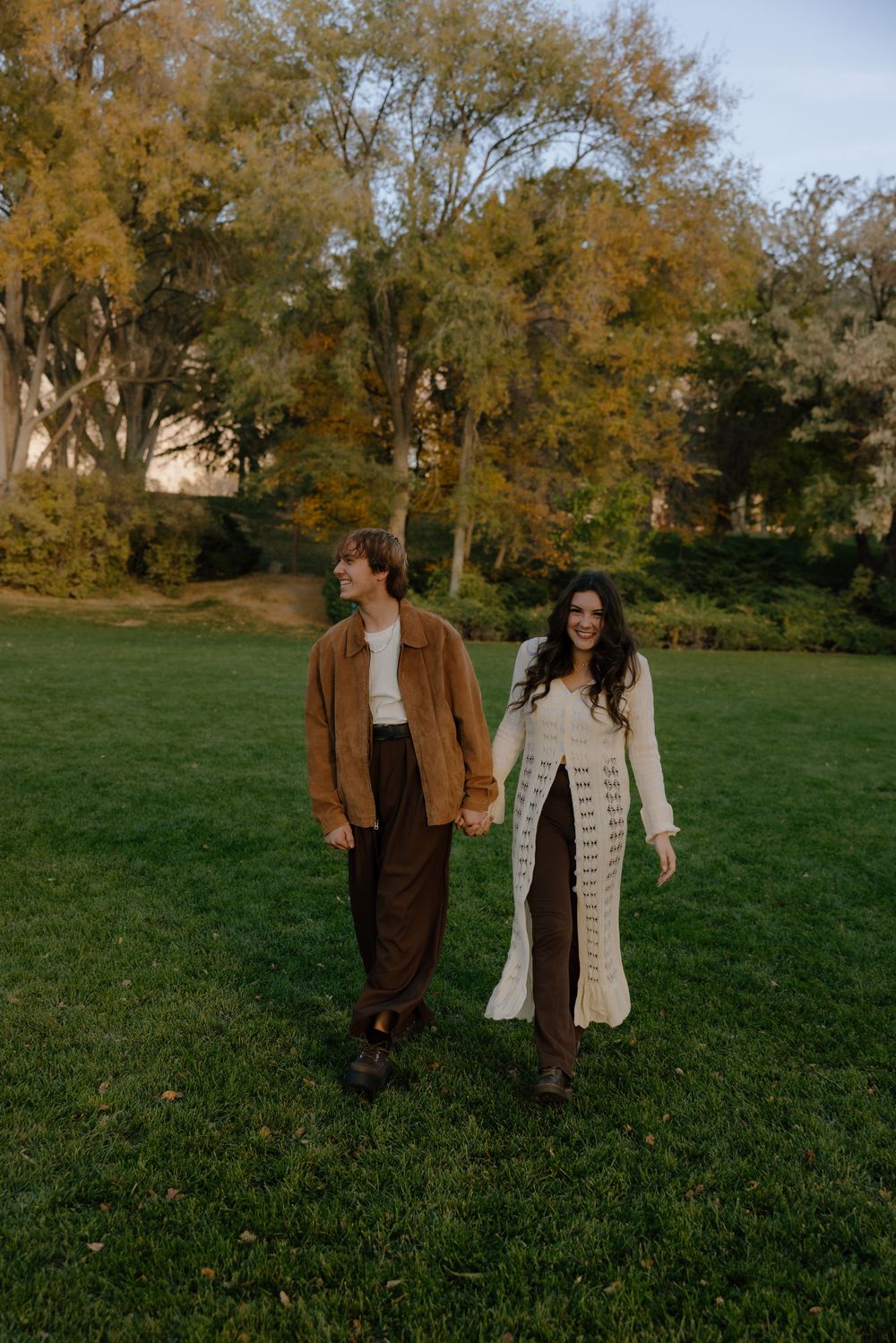 A couple walks together on a grassy field during autumn with trees in the background.