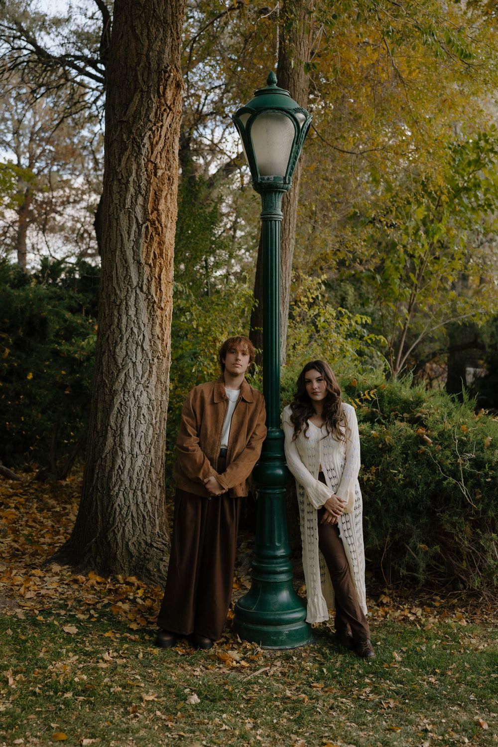 Two people in autumn outerwear stand near a vintage green lamppost in a park with fallen leaves and pine trees.