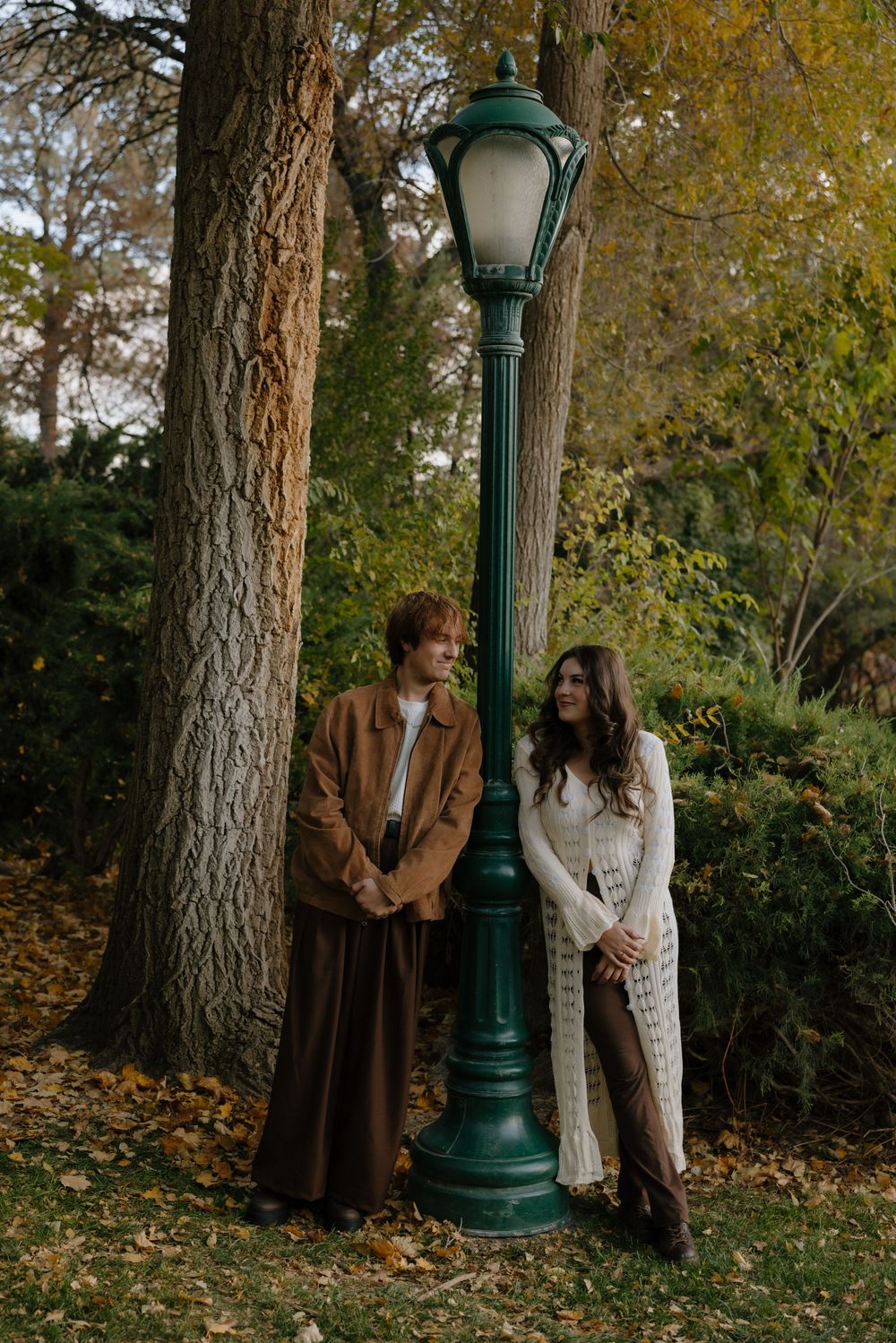 A romantic autumn scene with a couple standing by a vintage green lamppost surrounded by fallen leaves in a park.