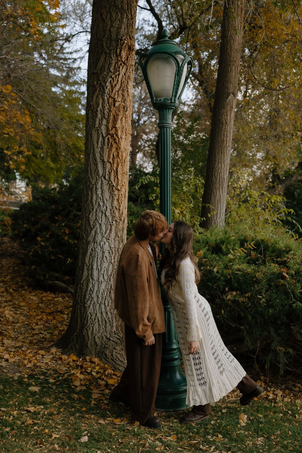 A romantic autumn scene with a couple by a vintage lamppost surrounded by fallen leaves and tall trees.