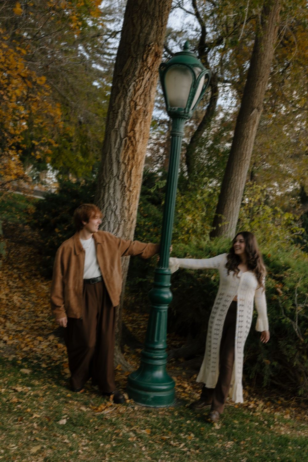 Two people in autumn attire stand near a vintage green lamppost surrounded by fall foliage in a park setting.
