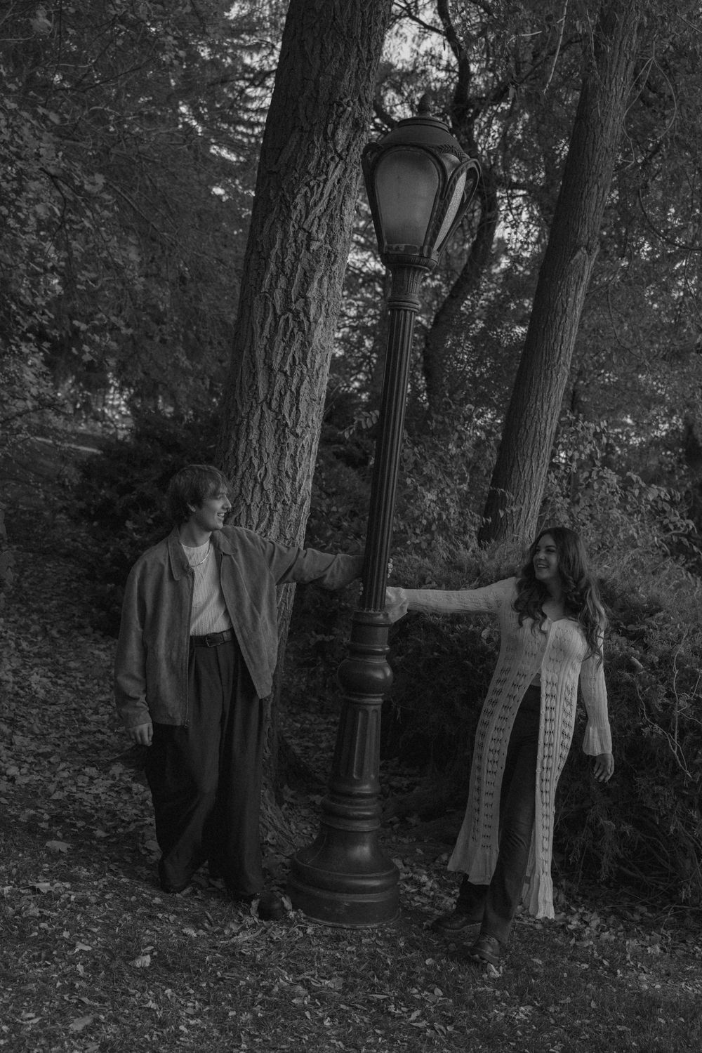 Two people stand near a vintage lamp post in a wooded area creating a moody black and white scene.