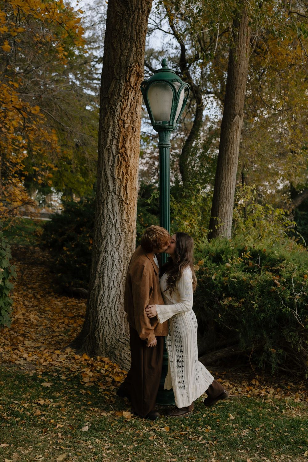 A vintage-inspired autumn scene with two people in coats standing beneath a green lamp post surrounded by fall foliage.