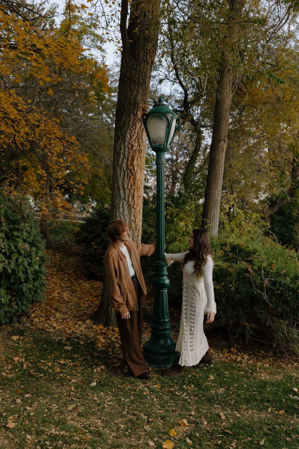 Two people walk through a wooded autumn scene with fallen leaves and tall trees with golden foliage.