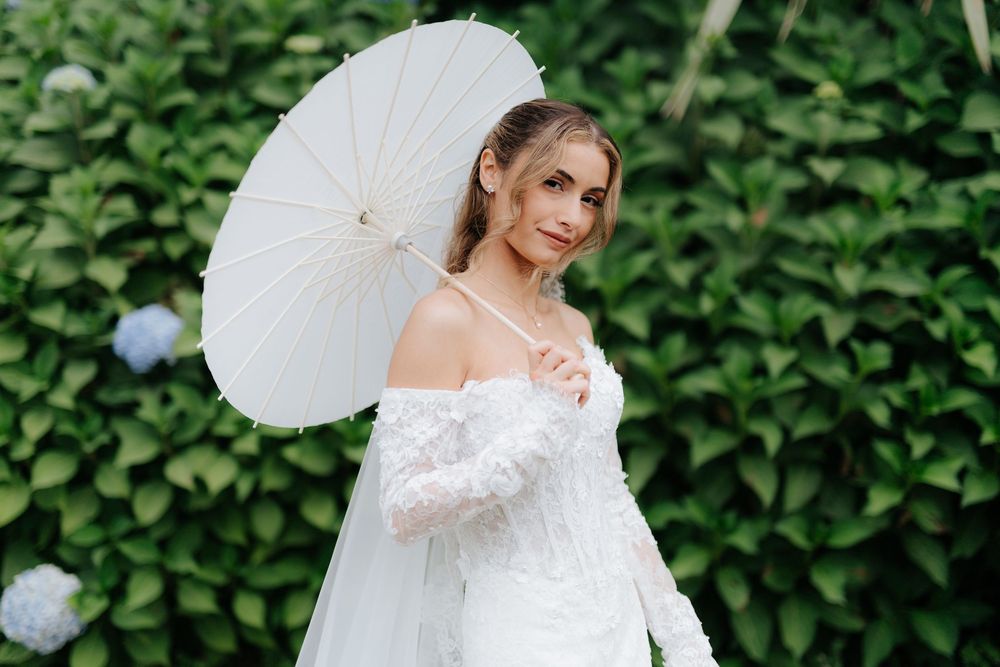 Bride in white lace gown holding a white parasol umbrella, posing outdoors near green hedges and hydrangeas.