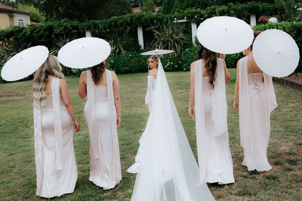 Bride in white gown and veil stands with bridesmaids holding white parasols in lush garden wedding setting.