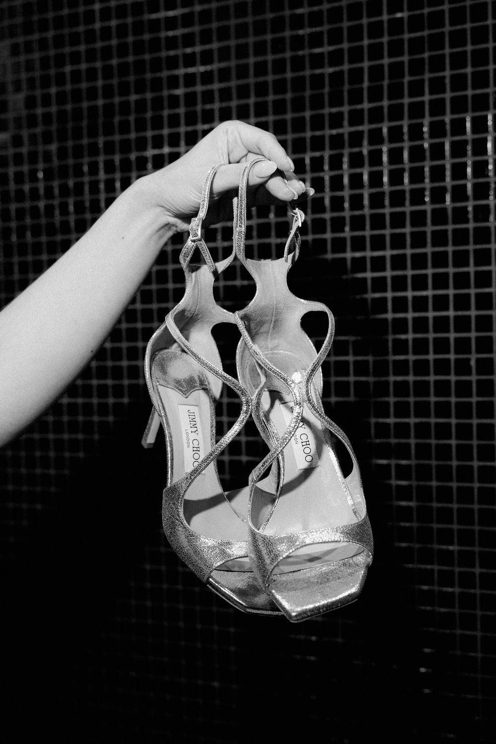 A hand holds strappy high-heel sandals by Jimmy Choo in a black and white photo.
